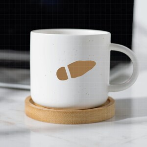 May include: A white ceramic mug with a brown footprint design, resting on a round bamboo coaster. The mug has a speckled texture and a curved handle. The footprint design is a light brown color.