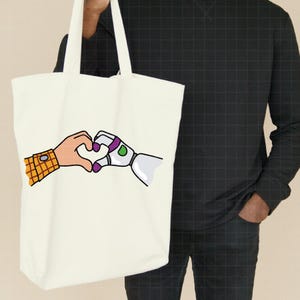 May include: Off-white tote bag featuring a heart shape formed by a cartoon hand and a robotic hand. The cartoon hand has a yellow and brown plaid sleeve. The robotic hand is white, purple, and green.