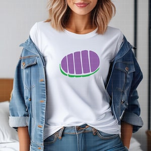 May include: White t-shirt featuring a purple and green graphic design. The design is an oval shape with vertical purple stripes and a green border. The shirt is worn with a blue denim jacket and blue jeans.
