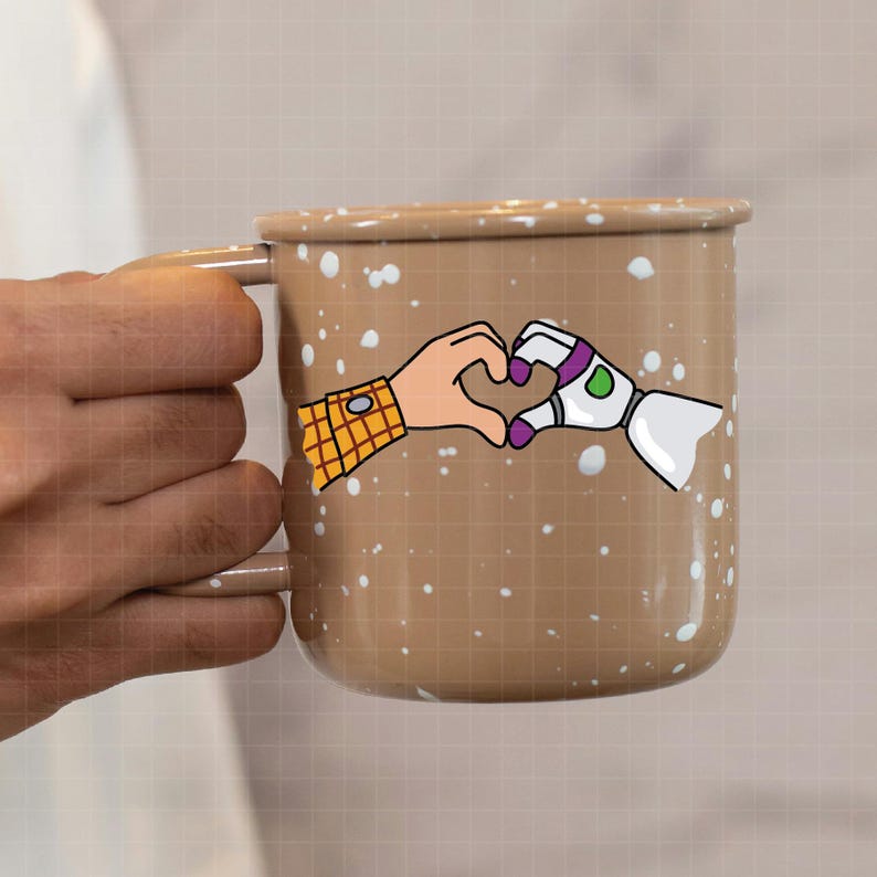 May include: A brown enamel mug with white speckles. The mug features a cartoon illustration of two hands forming a heart shape. One hand is a human hand, and the other is a robot hand. The mug is being held by a person.