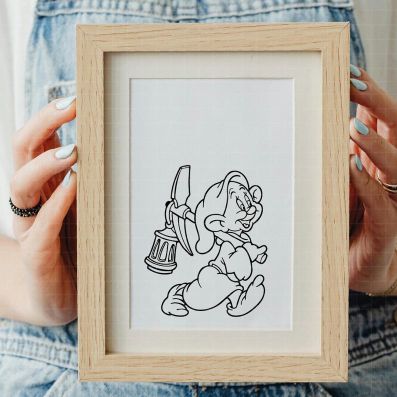 Puede incluir: Ilustraci&oacute;n en blanco y negro enmarcada de un personaje de dibujos animados sosteniendo un pico y una linterna. La obra de arte est&aacute; en un marco de madera clara, sostenida por una persona con un peto vaquero azul.