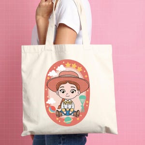 May include: A cream-colored tote bag featuring a cartoon illustration of a cowgirl character. The illustration is set within an oval frame with stars, clouds, and a rainbow. The cowgirl wears a hat, a yellow shirt, and blue jeans.