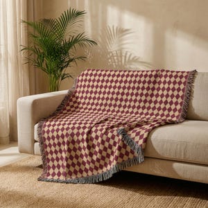 May include: A woven throw blanket featuring a repeating diamond pattern in deep burgundy and cream colors. The blanket has a textured finish and is finished with frayed, fringed edges along the sides, draped over a neutral-toned sofa.