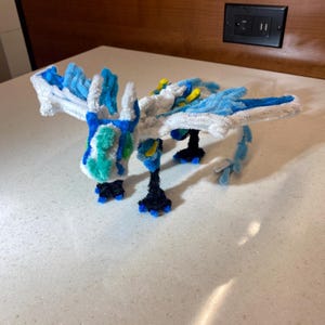 May include: A handcrafted pipe cleaner dragon sculpture. The dragon has a white body with blue and turquoise accents on its wings, head, and tail. The dragon's feet are black with blue claws, and it has yellow details.