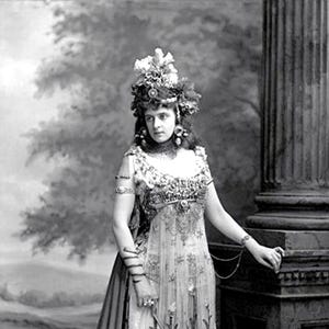 May include: A vintage black and white photograph of a woman in an elaborate dress and headdress. The dress is adorned with intricate details, and the headdress includes floral and decorative elements. The woman is positioned near a classical column.
