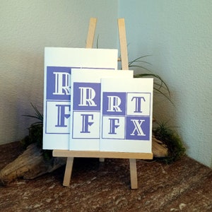 May include: Three white cards with a purple border and white letters on a wooden easel. The cards are different sizes and each has a different letter: R, F, T, and X.