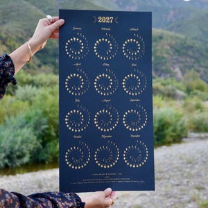 May include: A dark blue 2027 wall calendar featuring gold foil moon phase illustrations arranged in spiral patterns for each month from January to December. The calendar includes text labels for each month and a legend at the bottom.