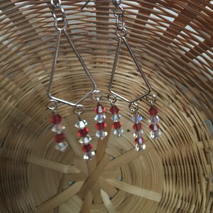 May include: A pair of silver dangle earrings with red and clear crystal beads hanging from a triangle-shaped wire.