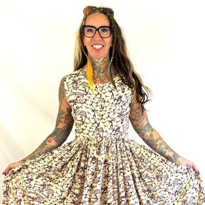 May include: A sleeveless, cream-colored dress with a brown print featuring huts, palm trees, and figures. The dress has a fitted bodice and a flared skirt. The fabric appears to be cotton or a similar lightweight material, suitable for warm weather.