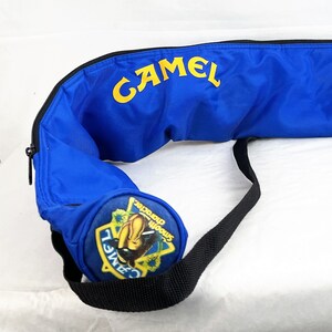 May include: A bright blue cylindrical carrying case with a black zipper and a black shoulder strap. The word CAMEL is printed in yellow on the side. A circular end cap features a yellow and blue logo with a camel graphic and the text Smooth character.