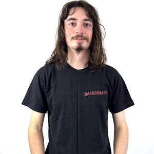 Peut inclure: Une personne portant un t-shirt noir avec le mot "BACKDRAFT" imprimé en rouge sur le devant. La personne porte un jean bleu.
