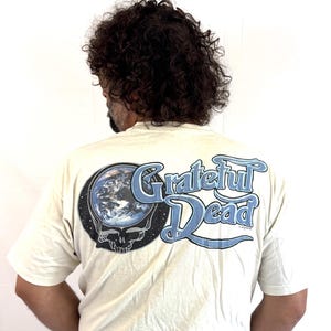 May include: Cream-colored t-shirt featuring the Grateful Dead logo on the back. The design includes a skull with a blue and white Earth graphic and the words "Grateful Dead" in a stylized font. The shirt is a classic band merchandise.