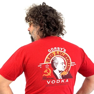 May include: Red t-shirt with a graphic featuring the text "Gorby's Red Lightning Vodka" and an image of a man's profile. The shirt also displays a hammer and sickle symbol and the words "100 Proofski".
