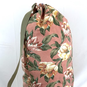 May include: A pink floral drawstring bag with a green shoulder strap. The bag is covered in a pattern of large, stylized flowers in shades of yellow, white, and pink, with green leaves.