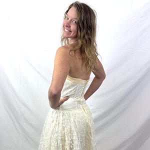May include: A strapless, ivory-colored dress with a lace overlay. The dress features a fitted bodice and a full, flowing skirt. The model is smiling and looking over their shoulder. The dress appears to be vintage or a formal occasion dress.