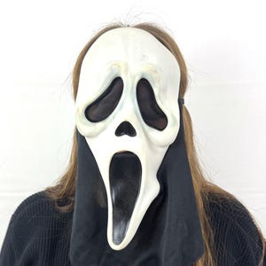 Peut inclure: Un masque de Scream, avec un visage blanc allongé, des orbites noires et une bouche béante. Le masque est attaché à une cagoule noire, créant une apparence fantomatique. La personne porte une chemise noire.