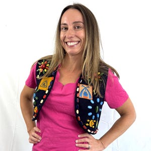 May include: A dark blue vest with colorful beaded embellishments and embroidered patches. The vest is worn over a pink t-shirt and blue jeans. The patches feature geometric and nature-inspired designs.
