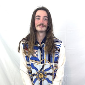 Puede incluir: Una camisa abotonada con estampado náutico. La camisa presenta un cuerpo color crema con mangas largas y cuello y puños azul marino. El panel frontal tiene un diseño de sol azul y dorado y motivos náuticos.