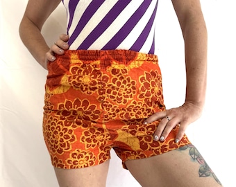 May include: Orange and yellow floral print shorts with an elastic waistband. The shorts are paired with a sleeveless top with diagonal purple and white stripes. The outfit is shown on a person.