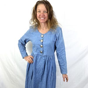 Pode incluir: Vestido comprido de ganga azul claro, com mangas compridas, decote redondo e botões prateados. O vestido tem cintura franzida e saia fluida, adequado para uso diário.