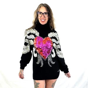 May include: A black turtleneck sweater with a large sequined heart design. The heart is pink and red with silver accents. The sweater has silver sequined wings on the shoulders.