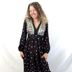 May include: A long, dark brown dress with a V-neck and long sleeves. The dress features a colourful paisley pattern in shades of red, blue, and teal. The dress has a fitted waist and a flowing skirt, creating a bohemian style.