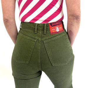 May include: Olive green high-waisted jeans with a classic five-pocket design. The jeans feature a Bongo brand patch on the back pocket. The model is wearing a pink and white striped top.
