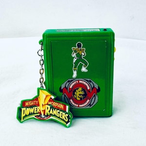 May include: Green plastic Power Rangers toy with a keychain. The toy features a green ranger character, the Mighty Morphin Power Rangers logo, and a red button. The keychain is attached to a silver chain.