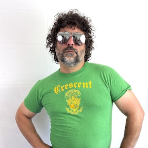 May include: A bright green short-sleeved t-shirt featuring the word Crescent in yellow gothic-style lettering above a yellow circular emblem with intricate details. The shirt is paired with dark blue denim jeans and reflective aviator sunglasses.