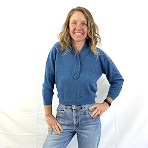 May include: A woman wearing a blue sweater and blue jeans. She is standing with her hands on her hips and smiling.