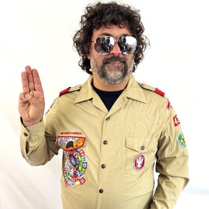 May include: A tan long-sleeve button-down shirt featuring Boy Scouts of America patches, red shoulder epaulets, and a collar. The shirt is worn with blue denim jeans and a brown belt with a metal buckle. The shirt displays various colorful embroidered patches.