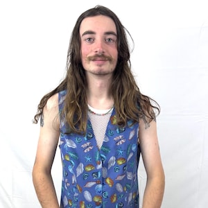 May include: A blue sleeveless vest adorned with a seashell design. The vest showcases a variety of seashells in white, orange, and teal hues. It is worn over a white mesh top and paired with brown trousers.