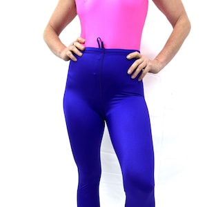 May include: A person wearing a pink leotard and blue leggings. The leggings are a solid, vibrant blue color. The leotard has a scoop neck and a drawstring. The person is standing with their hands on their hips.