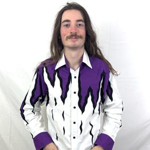 Puede incluir: Camisa blanca abotonada con un diseño llamativo con patrones de llamas moradas y negras en los hombros y la parte superior del pecho. La camisa tiene un cuello y puños clásicos en un color morado a juego. La camisa se combina con vaqueros azul oscuro.