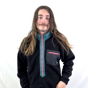 May include: Black fleece pullover with a teal collar and a red zipper. The pullover has a black pocket with a red trim. The model is wearing olive green cargo pants.