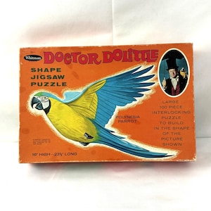 May include: A 100-piece jigsaw puzzle featuring a colorful parrot with the text "Doctor Dolittle" and "Polynesia Parrot" on the box. The puzzle is 16 inches high and 23 1/2 inches long.