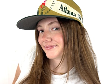 Puede incluir: Gorra de béisbol vintage de los Juegos Olímpicos de Atlanta 1996. La gorra es de color verde oliva con una visera gris oscuro y presenta el logotipo y el texto de Atlanta 1996. Tiene una banda blanca con la palabra "Atlanta" impresa.