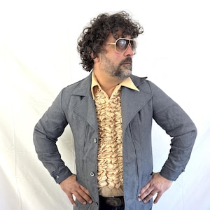May include: A gray jacket with a notched collar and button closure. The jacket is worn over a ruffled beige shirt and a yellow collared shirt. The person is wearing blue jeans and aviator sunglasses.