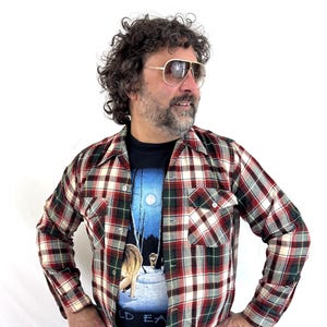 May include: A long-sleeve button-up shirt featuring a plaid pattern in red, green, white, and black. The shirt has two chest pockets with button flaps, a pointed collar, and a front button closure. It is worn over a black t-shirt with a graphic print.