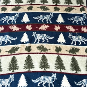 May include: A rectangular fleece blanket with a repeating pattern of wolves, pine trees, and leaf clusters. The blanket features a navy blue border and stripes of navy blue, burgundy, beige, and olive green.