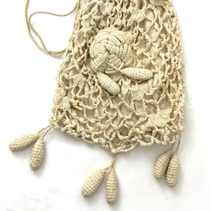 May include: Cream-colored crocheted drawstring bag with a floral design and dangling tassels. The bag features a delicate openwork pattern and a woven cord for closure. The bag is approximately 8 inches tall.