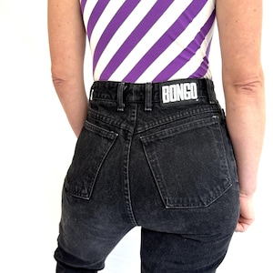 Puede incluir: Vaqueros negros de mezclilla con cintura alta y la marca "BONGO" en la cinturilla trasera. La parte superior es un body sin mangas con rayas diagonales blancas y moradas.
