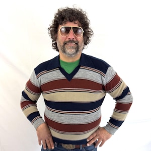 May include: A long-sleeve V-neck sweater featuring horizontal stripes in navy blue, rust red, light gray, and beige. The sweater has a ribbed collar and cuffs, worn over a solid green shirt, paired with blue denim jeans and gold-rimmed sunglasses.