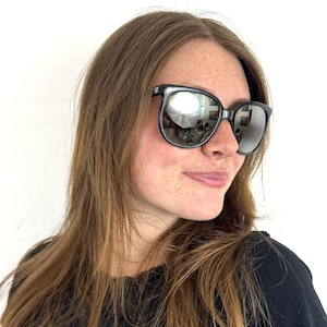 May include: Black sunglasses with rounded frames and reflective silver lenses. The frames feature a small white logo on the upper corner of the temple. The lenses provide a mirror-like finish that reflects the surrounding environment clearly.