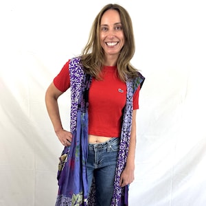 May include: A sleeveless, long patchwork vest in shades of purple, blue, and green, featuring floral and patterned designs. The vest is worn over a red short-sleeved shirt and blue jeans.