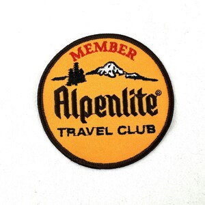 Könnte beinhalten: Ein runder, orangefarbener Patch mit braunem Rand. Der Patch trägt den Text "MEMBER" in Rot, "Alpenlite" in Schwarz und "TRAVEL CLUB" in Schwarz. Im Hintergrund sind ein weißer Berg und ein schwarzer Baum zu sehen.