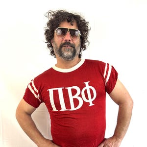 Könnte beinhalten: Rotes Ringer-T-Shirt mit weißen Streifen auf den Ärmeln und den griechischen Buchstaben ΠΒΦ in Weiß. Das Shirt hat einen weißen Kragen und wird mit einer blauen Jeans kombiniert. Die Person auf dem Bild trägt eine Pilotenbrille.