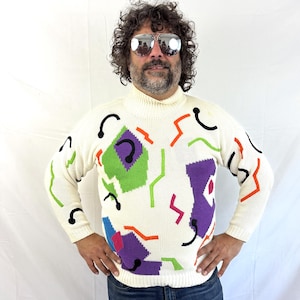 May include: A cream-colored turtleneck sweater featuring a knit pattern of abstract shapes in bright purple, green, orange, and black. The design includes scattered geometric lines and solid blocks of color across the front and sleeves.
