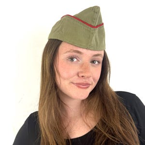 May include: An olive green military-style cap with a red trim. The cap has a triangular shape and is made of a textured fabric. A vintage style cap, it is a classic military accessory.