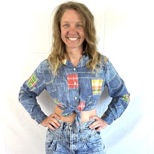 May include: A denim shirt with colorful plaid patches is tied at the waist, paired with acid wash jeans. The shirt has long sleeves and a collared neckline. The jeans have a high waist and a tapered leg. The outfit is a vintage style.